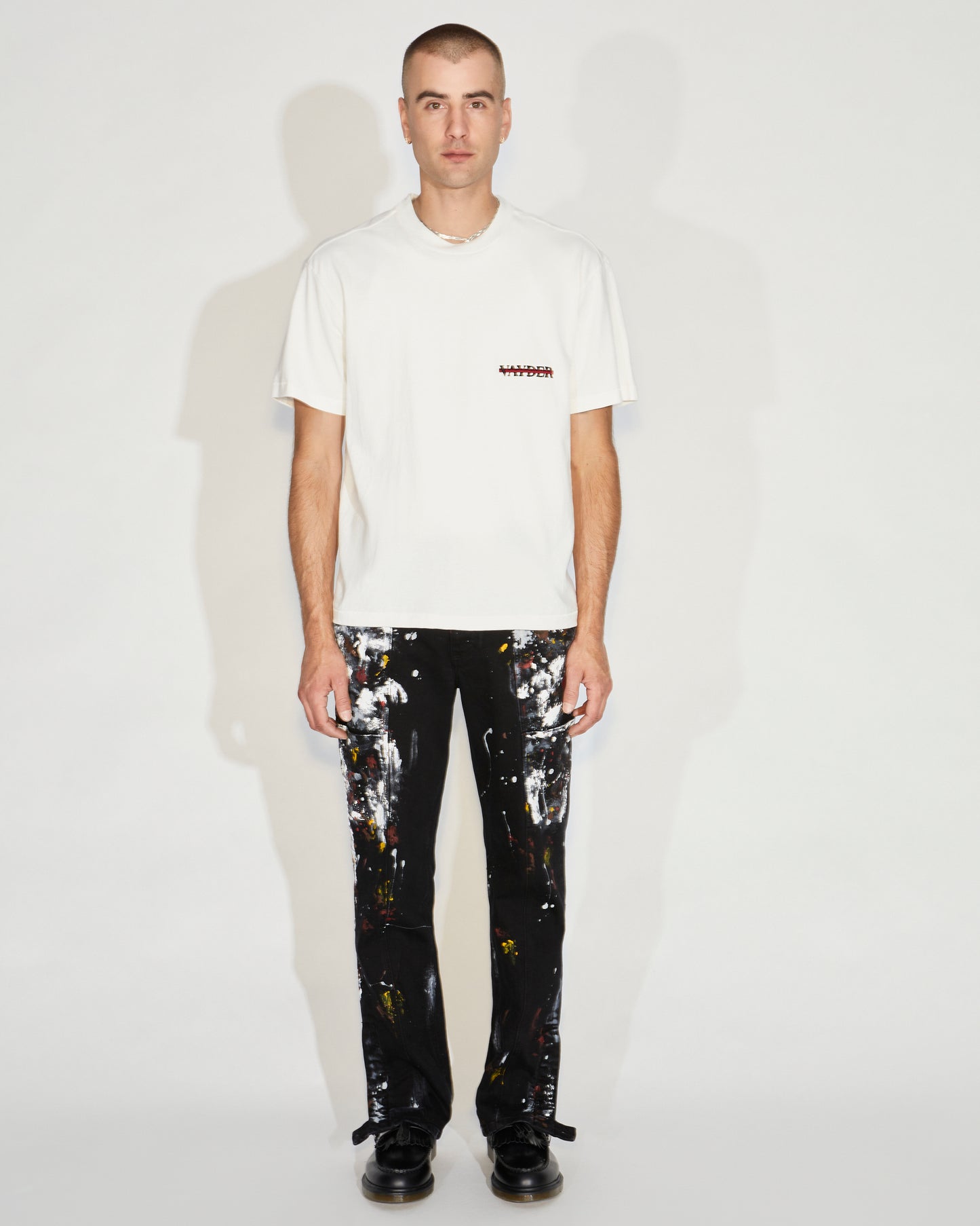 Man standing wearing black pants with paint splatter and a white tee and black shoes
