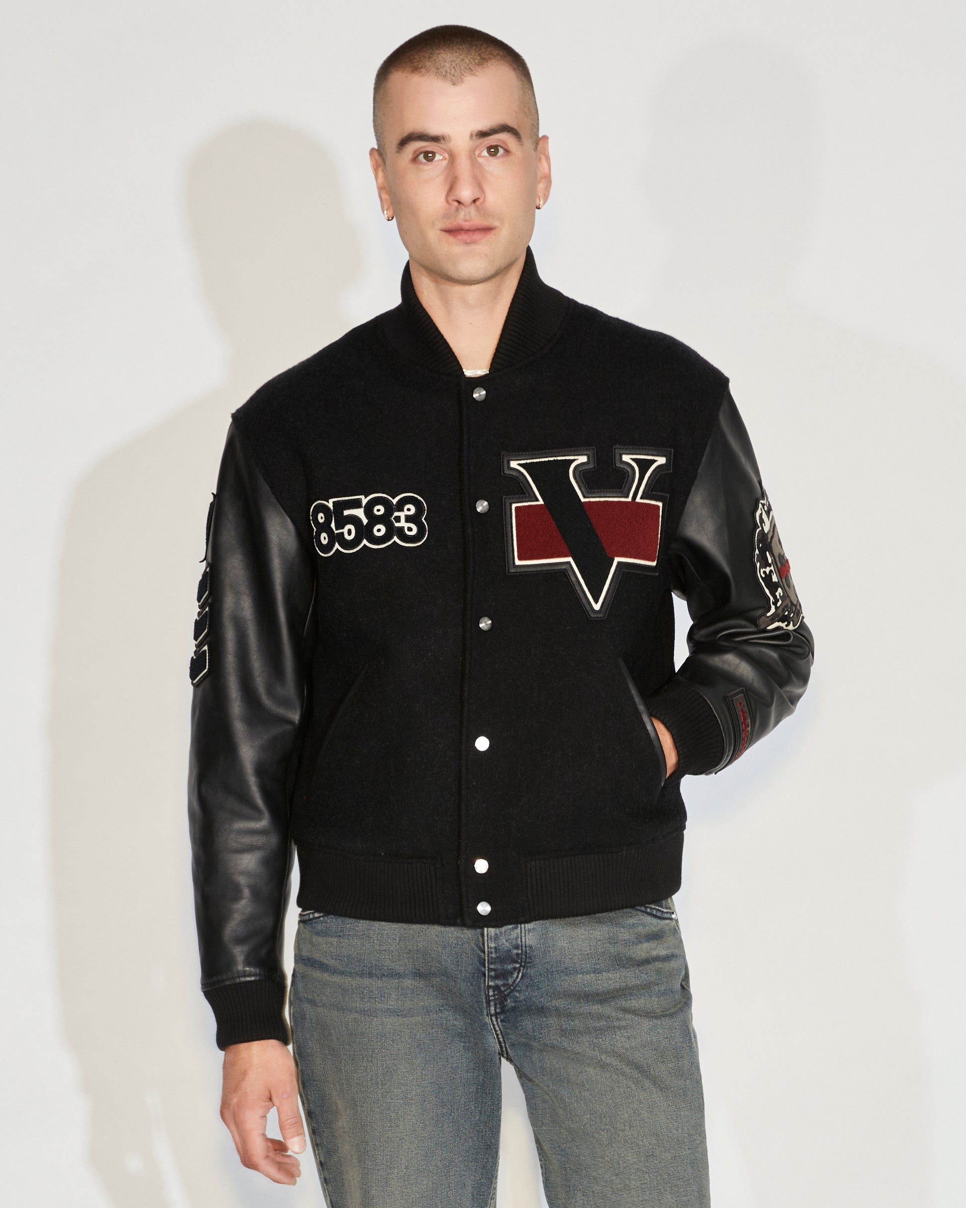 Man standing wearing a black button-up varsity jacket with chest with arm patches and jeans